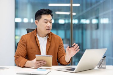 Başarılı bir işadamı Webinar eğitimi alırken, iş yerinde ofis içinde not defterine veri yazarken, Asyalı bir adam çevrimiçi konferans için dizüstü bilgisayar kullanıyor..