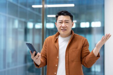 Turuncu tişört giyen Asyalı bir adam ofisteki cep telefonunda okudukları yüzünden kafası karışmış gibi görünüyor..