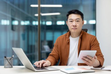 Asyalı bir işadamı aynı anda hem bilgisayarı üzerinde çalışırken hem de ofiste sunum için tablet kullanıyor..