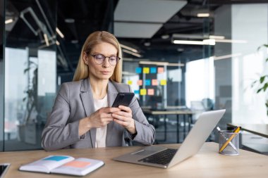 Ceketli bir iş kadını modern ofis ortamında bir masada otururken cep telefonunu kullanıyor..