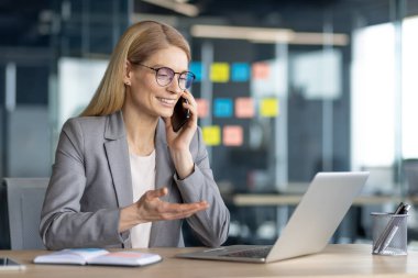 Gözlük takan gülümseyen bir iş kadını ofisinde dizüstü bilgisayarla otururken telefonla konuşuyor..