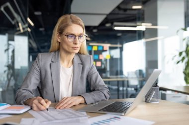 Sarışın, profesyonel bir kadın belgelere bakıyor ve bilgisayarını modern ofis ortamında kullanıyor..