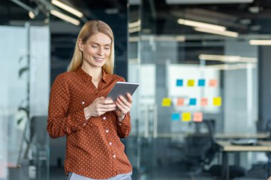 Sarışın bir iş kadını, modern bir ofiste cam duvarlar ve renkli yapışkan notlarla dolu bir tablet kullanıyor..