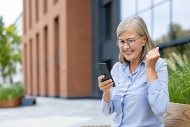 Heyecanlı, gözlüklü olgun bir kadın, güneşli bir günde dışarıda bankta oturmuş akıllı telefonuna bakarken kutlama yapıyor..