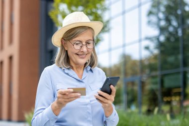 Gözlüklü ve şapkalı gülümseyen bir son sınıf öğrencisi şehir ortamında kredi kartıyla alışveriş yapıyor..