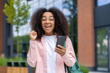 Afro saçlı genç bir kadın güneşli bir günde dışarıda sırt çantasıyla telefonuna bakıyor..