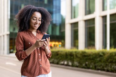 Genç bir kadın ofis binasının önünde akıllı telefonunu kontrol ediyor, modern teknolojinin tadını çıkarıyor ve bağlantıda kalıyor..