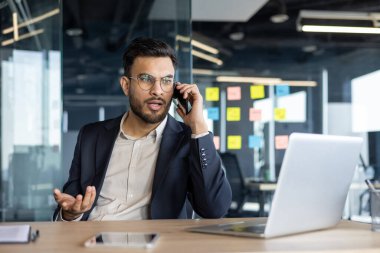 Ciddi ve odaklanmış bir adam ofisin içinde telefonla konuşuyor. İş adamı takım elbiseli. Masasında dizüstü bilgisayarla oturuyor..