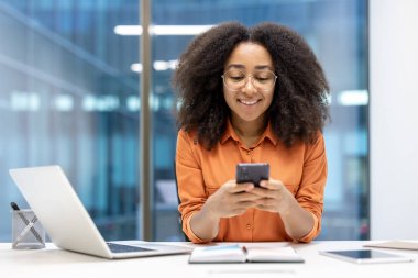 Başarılı neşeli kadın telefonunda başvuru kullanıyor. İş kadını iş yerinde elinde akıllı telefonuyla oturuyor, mesaj yazıyor ve sosyal ağlara takılıyor. Ofisteki mutlu bayan çalışan.