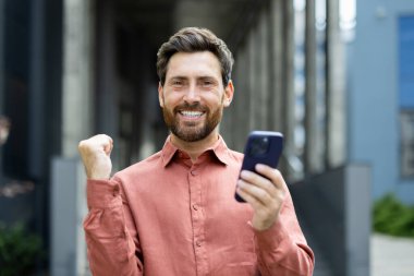 Düğmeli tişört giyen sakallı bir adam elinde akıllı bir telefon tutuyor ve başarısını kutluyor..