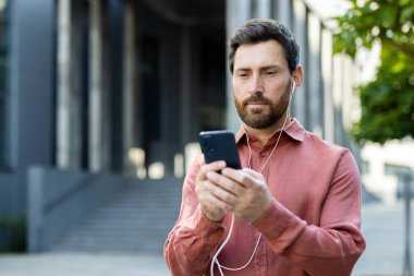 Sakallı bir adam modern bir binanın önünde duruyor, müzik dinliyor, güneşli bir günde cep telefonuna bakıyor..
