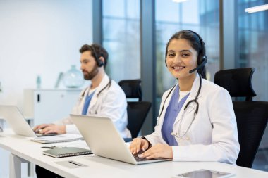 Kulaklık takan gülümseyen genç bir bayan doktor ve steteskop arka planda erkek bir meslektaşıyla birlikte bilgisayarında çalışıyor..