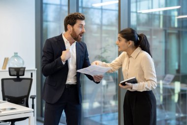 Bir adam ve bir kadın ofiste ateşli tartışmalar yapıyorlar. Belgeleri tutarken, işyeri çatışmalarını gösterirken.