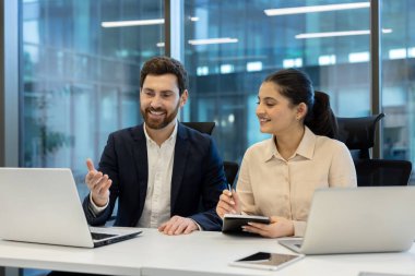 Bir erkek ve bir kadın bir ofis ortamında birlikte çalışıyorlar, dizüstü bilgisayarlarını kullanarak bir proje üzerinde çalışıyorlar ve notlar alıyorlar..