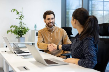 Bir erkek ve bir kadın, modern ofis ortamında dizüstü bilgisayarlarla el sıkışıyor, iş anlaşmasını ya da ortaklığı simgeliyor..