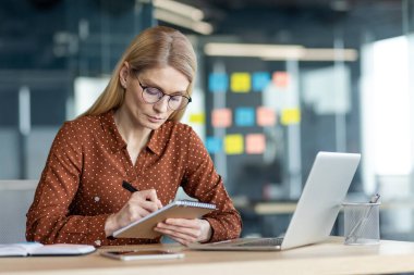 Ofisteki masada otururken not defterine veri yazmayı düşünen ciddi bir kadın. Çevrimiçi iş toplantısındaki iş kadını. Eğitimsel webinar izleme.