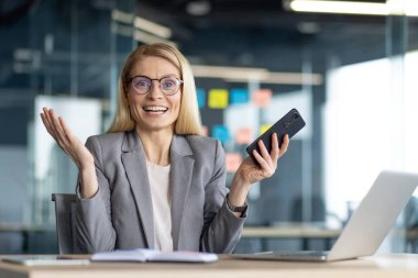 Heyecanlı bir iş kadını akıllı telefonunu tutarak başarıyı kutluyor, modern bir ofis ortamında masasında çalışıyor..