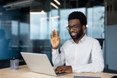Modern bir ofisteki bilgisayarında iş arkadaşları ya da müşterileriyle bağlantı kuran gözlüklü, gülümseyen bir Afrikalı Amerikalı adam..