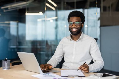 Mutlu, genç bir profesyonel dizüstü bilgisayarda çalışır, notlar alır ve masasında cam duvarlı verileri gözden geçirir..