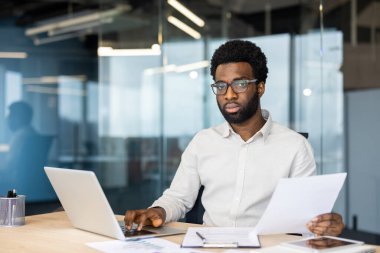 Belgeleri inceleyen ve modern ofis ortamında dizüstü bilgisayarı üzerinde çalışan odaklanmış profesyonel dengeler, üretkenlik ve çalışkanlık sergiliyor.
