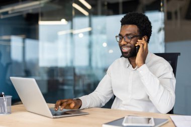 Gözlüklü neşeli bir Afro-Amerikalı, modern ofis ortamında cep telefonuyla konuşurken dizüstü bilgisayarında çalışıyor..