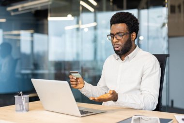 Kafası karışmış gözlüklü bir Afrikalı Amerikalı modern bir ofiste kredi kartı ve laptopunu kullanarak online işlem yapmaya çalışırken sorunlarla karşılaşır..