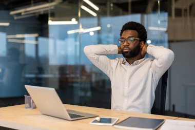 Gözlüklü profesyonel bir adam ellerini boynunun arkasına koyup modern ofis ortamında mola veriyor..