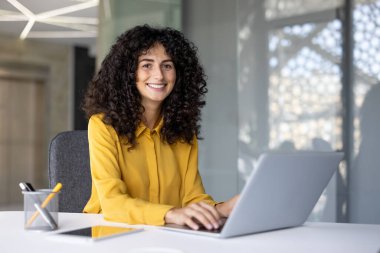 Ofisteki iş yerinde başarılı bir iş kadınının portresi. Kadın gülümsüyor ve kameraya bakıyor, dizüstü bilgisayarın klavyesinde yazıyor..