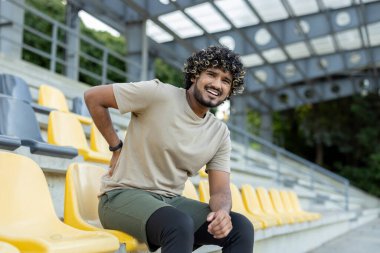 Genç atletik adam stadyum koltuklarında otururken acı içinde kıvranıyor. Açık hava antrenmanı sırasında sırt ağrısı çekiyor..