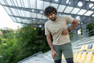 Stadyumda fiziksel bir egzersiz sırasında yan ve dizini tutan genç sporcu rahatsızlık ya da acı çekiyor..