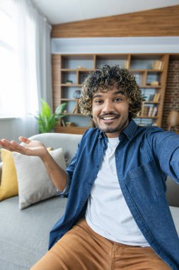 Telefon webcam görüntüsü. Adam gülümsüyor ve kameraya bakıyor, iş arkadaşları ve arkadaşlarıyla konuşuyor akıllı telefon uygulaması kullanıyor, oturma odasında kanepede oturuyor..
