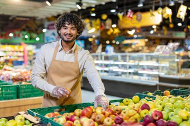 Dost canlısı bir market çalışanı kameraya gülümserken bir elma tutuyor, taze ürünlerle çevrili..