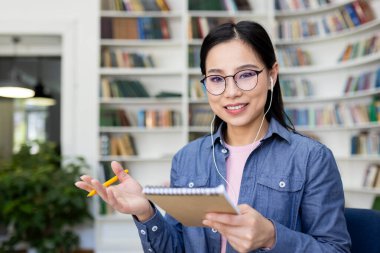 Kütüphanede gözlüklü gülümseyen Asyalı kadın, kulaklık takıyor, not defteri tutuyor, video görüşmesi sırasında el kol hareketi yapıyor..