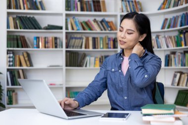 Masada oturan Asyalı bir kadın, kütüphanede dizüstü bilgisayar kullanıyor, boyun ağrısı çekiyor, arka planda kitaplıklar var..