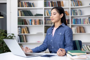 Huzurlu bir Asyalı kadın kütüphane ortamında meditasyon yapıyor, işini ve iyiliğini farkındalıkla dengeliyor. Yakınlarda bir laptop ve tablet vardı..
