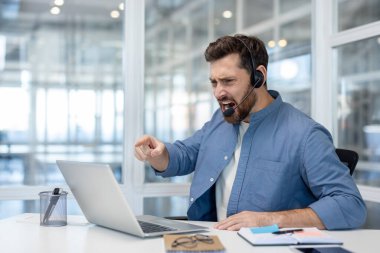 Kulaklık takan öfkeli bir müşteri hizmetleri temsilcisi dizüstü bilgisayarındaki bir soruna tepki olarak bir patlama yaşıyor. Hoşnutsuz bir ifadeyle işaret edip bağırıyor..