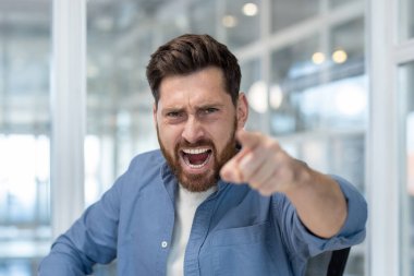 Mavi gömlekli kızgın bir adam doğrudan izleyiciyi işaret ediyor, ağzı açık bağırıyor ve sert bir ifade takınıyor..