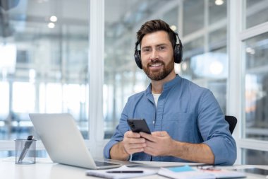 Kulaklık takan gülümseyen bir işadamı ofis ortamında kameraya bakarak bir akıllı telefon tutuyor. Rahatlamış ve işine dalmış görünüyor..
