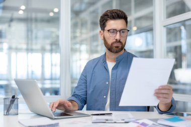 Bir işadamı masasında dizüstü bilgisayarı üzerinde çalışırken belgelere göz atar, modern bir ofiste analiz ve planlama üzerine odaklanır..