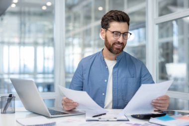 Gülümseyen profesyonel bir adam masasında belgelerini gözden geçiriyor. Parlak bir ofis ortamında, işle meşgul. O, gözlük ve mavi bir gömlek giyiyor..