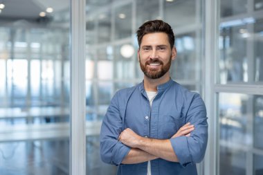 Çapraz kollu neşeli bir işadamı, modern, cam duvarlı bir ofiste, parlak bir şekilde gülümsüyor..