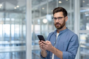 Sakallı, gözlüklü ve mavi gömlekli odaklanmış bir adam akıllı telefonunu tutarken doğrudan kameraya bakıyor..
