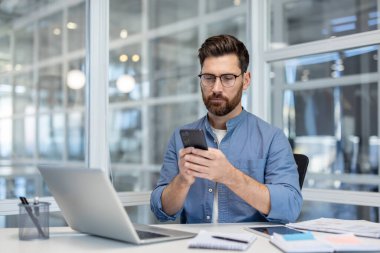 Gözlüklü bir işadamı masasında akıllı telefonunu kullanıyor. Yanında dizüstü bilgisayar ve belgeler var. Modern ofis ayarları pencerelerden görülebiliyor..