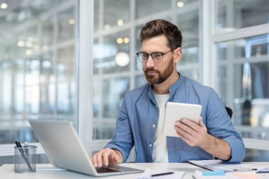 Odaklanmış bir profesyonel, elinde tablet tutarken bir dizüstü bilgisayarla modern ofis teknolojisini sergiliyor. Adam gözlük takıyor..