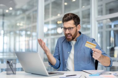 Modern bir ofiste, elinde kredi kartıyla bilgisayarına mutsuz bir ifadeyle bakan sinirli bir işadamı..