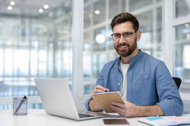 Modern ofis ortamında çalışan sakallı ve gözlüklü gülümseyen bir adam not defterine odaklanmış. Etrafı dizüstü bilgisayar ve diğer ofis malzemeleriyle çevrili..