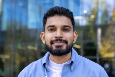 Sakallı yakışıklı bir adam dışarıda dururken doğrudan kameraya gülümsüyor. Arka plan cam pencereli modern bir binayı gösteriyor..