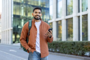Sırt çantalı gülümseyen bir adam akıllı telefonunu modern bir binanın dışında tutuyor ve şehrin güneşli gününün tadını çıkarıyor..