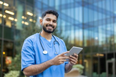Önlüklü bir Hintli doktor elinde tablet tutarken gülümsüyor, muhtemelen dışarıda binanın arka planındaki hasta verilerini inceliyor..
