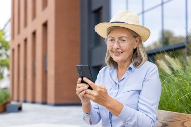 Şapkalı ve gözlüklü yaşlı bir kadın dışarıda akıllı telefon kullanırken gülümsüyor, havanın tadını çıkarıyor..